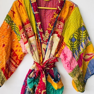 May include: A colorful patchwork robe with a tie belt, hanging on a yellow hanger. The robe features a variety of patterns and colors, including red, yellow, green, and pink. The robe is made of a quilted fabric.
