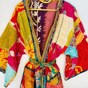 Puede incluir: Una vibrante bata kimono patchwork en una variedad de colores, incluyendo rojo, amarillo, rosa y azul. La bata tiene un cinturón y está hecha de tela acolchada. La bata está colgada en una percha amarilla.