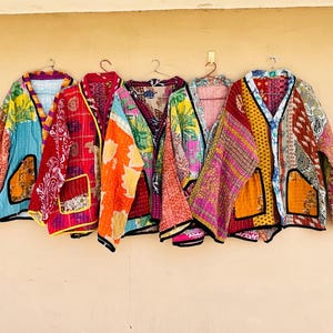 May include: A row of colorful, patchwork jackets hanging against a beige wall. Each jacket features a unique combination of patterns and colors, including red, orange, blue, and yellow. The jackets have a V-neck design and long sleeves.