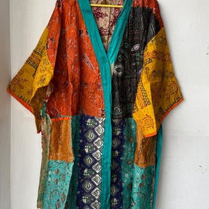 May include: A patchwork kimono in vibrant colors, including orange, yellow, teal, and navy. The kimono features a variety of patterned fabrics and a teal trim along the edges. It is hanging on a yellow hanger.