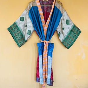 May include: A colorful kimono robe with a mix of patterns and colors, including blue, orange, green, and red. The robe is hanging on a hanger against a yellow background. The robe has a belt.