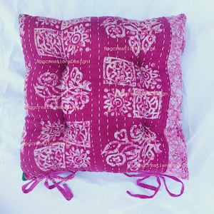 May include: A pink and white patterned cushion with a square shape and a button in the centre. The cushion has a white border and is tied with pink ribbons.
