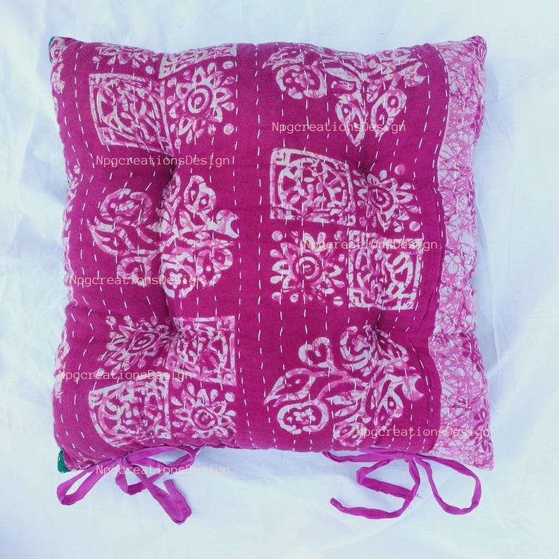 May include: A pink and white patterned cushion with a square shape and a button in the centre. The cushion has a white border and is tied with pink ribbons.