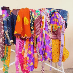 May include: A collection of colorful, patterned scarves hanging on a white metal rack. The scarves display a variety of vibrant colors, including orange, purple, pink, and yellow, with floral and leaf designs. The fabrics appear to be lightweight.