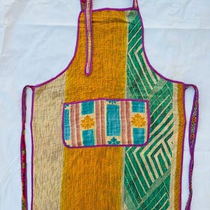 May include: A patchwork apron with a variety of colors and patterns. The apron features a large pocket with a floral design and is trimmed with a purple border. The apron has a neck strap and waist ties.