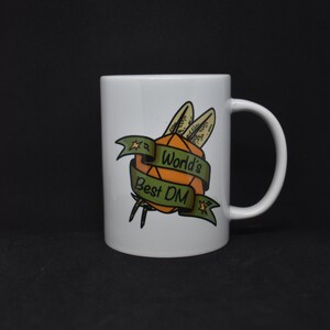 Puede incluir: Taza de cerámica blanca con un gráfico verde y naranja de un dado con alas y una pancarta que dice "World's Best DM".