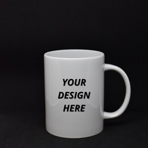 Könnte beinhalten: Eine weiße Keramiktasse mit einer schwarzen Aufschrift, die "YOUR DESIGN HERE" lautet.