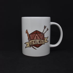 Può includere: Tazza in ceramica bianca con un'illustrazione marrone e bianca di un dado con un solo punto, due frecce e un pugnale. Il testo "Just Roll With It" è su uno stendardo giallo.