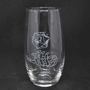 Puede incluir: Un vaso de vidrio transparente con un diseño de una mano esquelética sosteniendo un dado único con el número uno en él.