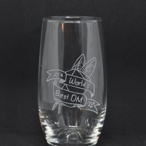 Könnte beinhalten: Ein klares Glas mit einem geätzten Design eines 20-seitigen Würfels, zwei Schwertern und einem Banner mit der Aufschrift "World's Best DM".