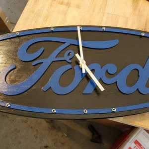 May include: An oval-shaped wall clock featuring the blue Ford script logo centered on a black background. The clock face is framed by a thin blue border accented with small silver-colored metal hardware pieces. It includes two silver clock hands.