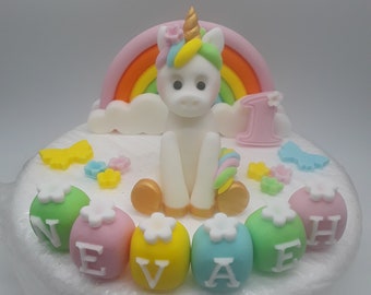 May include: A white fondant cake decorated with a unicorn, rainbow, and clouds. The cake is also decorated with colorful letters spelling out the name "NEVAEH".
