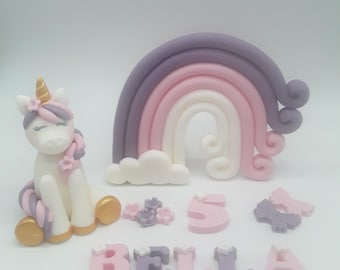 May include: A white unicorn cake topper with a pink and purple rainbow, clouds, flowers, and the name "BELLA" and the number "5" in pink and purple.
