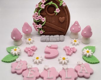 May include: A pink and brown fairy door cake topper with a number 5 and the name "BELLA" spelled out in pink letters. The topper also includes pink mushrooms, white daisies, and green leaves.