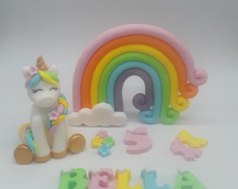 May include: A white unicorn cake topper with a rainbow, a cloud, flowers, the number 5, and the name "BELLA" in colorful letters.