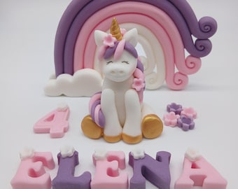 May include: A fondant cake topper set featuring a white unicorn with a gold horn, purple and pink mane, and floral accents. Behind it is a purple, pink, and white rainbow. Includes the number 4 and the name ELENA in pink and purple letters.