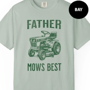 May include: A light green t-shirt with the words "FATHER MOWS BEST" printed in green. The graphic features a vintage lawn tractor. The shirt is a crew neck and has short sleeves.