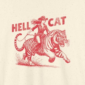 May include: A cream-coloured t-shirt with a red graphic of a woman riding a tiger. The text "HELLCAT" is printed in red above the graphic.