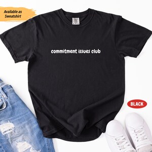 Puede incluir: Camiseta negra con el texto blanco "commitment issues club". La imagen muestra la camiseta con vaqueros azules y zapatillas blancas. Un pequeño rectángulo naranja indica que el artículo también está disponible como sudadera.