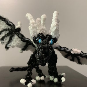 May include: A black and white rubber band dragon with blue eyes. The dragon has a white crown and wings.