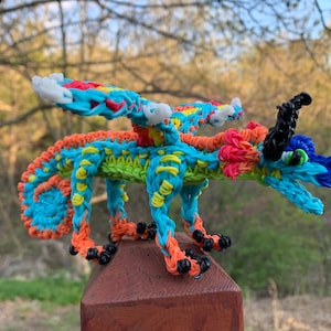 May include: A colorful rainbow loom dragon with blue, green, orange, yellow, and red bands. The dragon has a black horn and white eyes. It is standing on a wooden post.