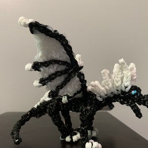 May include: A black and white rubber band dragon with a white mane and wings. The dragon has blue eyes and is posed in a standing position with its wings spread.
