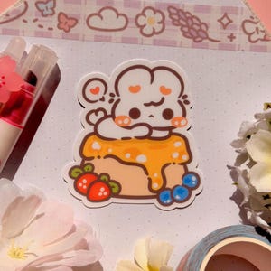May include: A die-cut sticker featuring a white cartoon rabbit resting on a yellow cake topped with orange glaze, red strawberries, and blue blueberries. The sticker is surrounded by a pink cherry blossom pen, white flowers, and patterned tape.