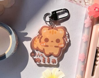 May include: A clear acrylic keychain featuring a cartoon orange cat with stripes and the text "olia miia". The keychain has a silver clasp and is surrounded by stationery items, including a pink craft knife and a floral pen.