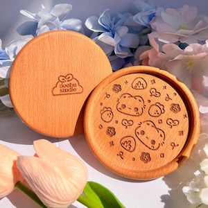 Könnte beinhalten: Zwei runde Untersetzer aus Holz. Auf einem Untersetzer ist der Text "doobu studio" eingraviert. Der andere Untersetzer hat ein eingraviertes Design von Cartoonfiguren, Blumen und anderen Formen. Die Untersetzer sind hellbraun.
