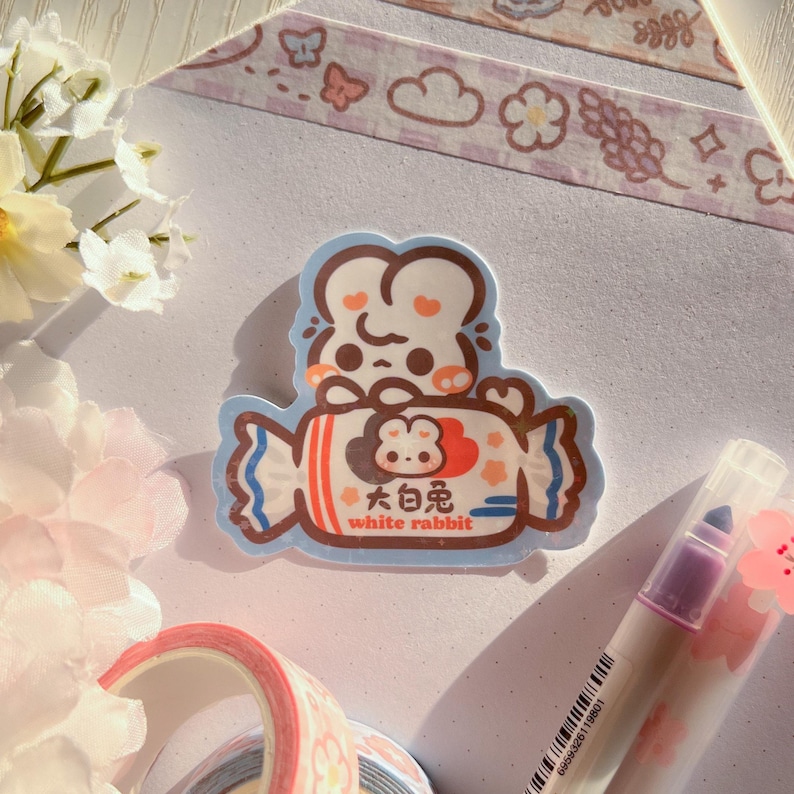 Puede incluir: Una pegatina con un conejo blanco de dibujos animados que sostiene un caramelo con el texto "大白兔 white rabbit" en &eacute;l. La pegatina est&aacute; sobre un fondo blanco con una cinta washi rosa y blanca.