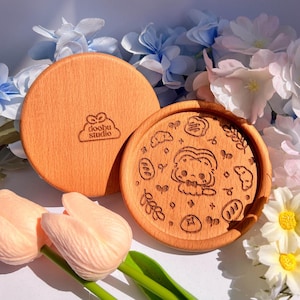 May include: Two round wooden coasters with a light brown finish. One coaster features a laser-etched design of a cartoon character and food items. The other coaster has the "doobu studio" logo. The coasters are surrounded by flowers.