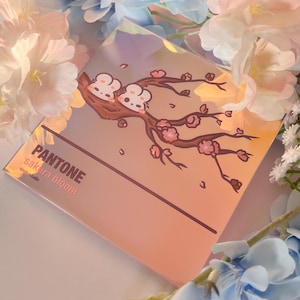 Puede incluir: Un espejo de bolsillo cuadrado, iridiscente y reflectante, con una ilustración de una rama de árbol marrón con dos pequeñas criaturas blancas y flores de cerezo rosas. El texto dice PANTONE sakura bloom y doobustudio en letras marrón oscuro.