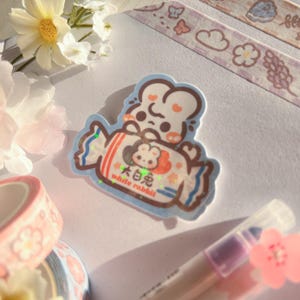 Puede incluir: Una pegatina azul y blanca con un conejo de dibujos animados sosteniendo una barra de caramelo. La barra de caramelo est&aacute; envuelta en papel de aluminio rojo y blanco y tiene el texto "白兔  white rabbit".
