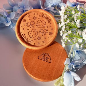 Puede incluir: Dos posavasos de madera con un acabado marrón claro. El posavasos superior presenta un diseño de nubes, lazos y flores. El posavasos inferior tiene el texto "doobu studio" en forma de nube.