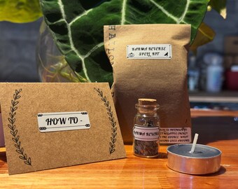 May include: A Karma Reverse Spell Kit with a brown paper bag, a small glass bottle, a tea light candle, and a card that says "HOW TO -" with a decorative border. The kit is on a wooden surface.