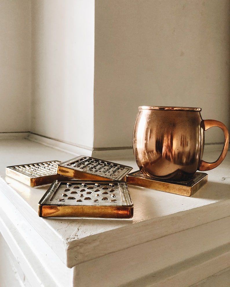 May include: Four gold-colored coasters with a geometric pattern and a copper mug on a white surface.