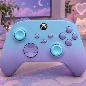 Puede incluir: Un mando de videojuegos inalámbrico con un degradado de color que va del azul claro en la parte superior a un morado suave en la inferior. Incluye dos joysticks azul claro, una cruceta morada y los botones estándar A, B, X e Y.