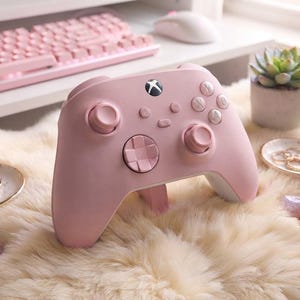 以下が含まれることがあります： 中央にXboxロゴボタン、2つのアナログスティック、方向パッド、A・B・X・Yと刻印された4つのアクションボタンを備えた、マットなパステルピンクのワイヤレスビデオゲームコントローラー。底面は白で、人間工学に基づいたグリップデザインが特徴です。