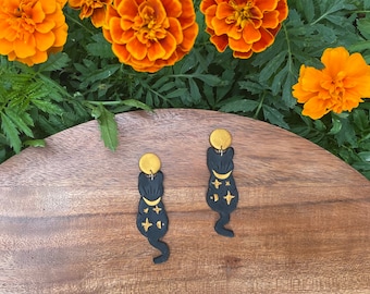 May include: A pair of black cat earrings with gold accents. The cats are adorned with a crescent moon and stars. The earrings are displayed on a wooden surface.