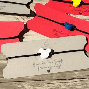 May include: A collection of bracelets with Mickey Mouse-shaped charms. The bracelets are black with charms in various colors, attached to gray and red cardstock tags. The tags have the text "You've been Pixie Dusted! Have a magical day!"