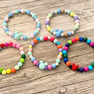 May include: Five colorful beaded bracelets. Each bracelet features a variety of pastel and vibrant colored beads. Some bracelets have a Mickey Mouse head design. The bracelets are displayed on a wooden surface.