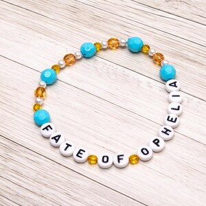 May include: A beaded bracelet with the words "FATE OF OPHELIA" spelled out in white letter beads. The bracelet features turquoise, gold, and pearl-colored beads. The bracelet is displayed on a light wood-grain surface.