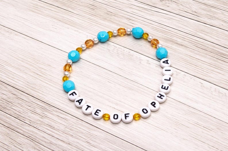 May include: A beaded bracelet with the words "FATE OF OPHELIA" spelled out in white letter beads. The bracelet features turquoise, gold, and pearl-colored beads. The bracelet is displayed on a light wood-grain surface.