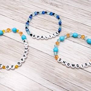 May include: Three beaded bracelets with white letter beads spelling "FATE OF OPHELIA", "OPALITE", and "SHOWGIRL". The bracelets feature turquoise, clear, and amber-colored beads. The bracelets are on a light wood-grain surface.