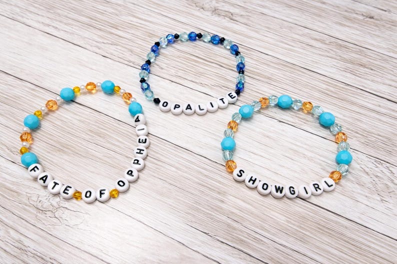 May include: Three beaded bracelets with white letter beads spelling "FATE OF OPHELIA", "OPALITE", and "SHOWGIRL". The bracelets feature turquoise, clear, and amber-colored beads. The bracelets are on a light wood-grain surface.