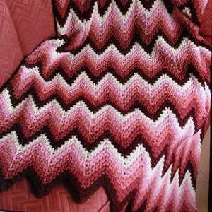 May include: A crocheted throw blanket with a chevron pattern in shades of pink, white, and brown. The blanket is draped over a red armchair.
