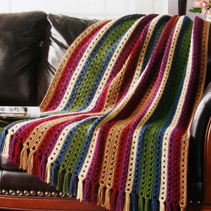 Op de afbeelding: Een meerkleurig gestreept gehaakt deken met franjes aan de randen. Het deken ligt op een bruine leren bank.
