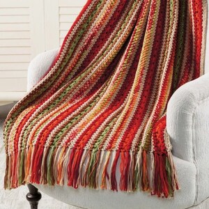 May include: A colorful, striped crochet throw blanket draped over a white armchair. The blanket features vertical stripes in shades of red, orange, green, and cream, with a fringed edge. The cozy blanket adds a warm, inviting touch to the room.