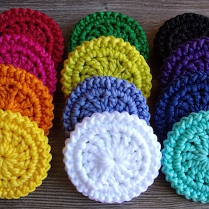 May include: A collection of handmade, circular crochet coasters in various vibrant colors, including red, orange, yellow, green, blue, purple, black, and turquoise. The coasters are arranged on a wooden surface, showcasing their textured, spiral design.