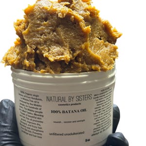 May include: A white jar of Natural by Sisters 100% Batana Oil. The jar contains a large, textured mass of brown oil. Text on the jar includes "nourish, recover and strength" and "unfiltered unadulterated". The jar is 8 oz.
